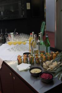 Ein Buffet mit Rotwein, Mineralwasser und Orangensaft in Flaschen und einigen gefüllten Gläsern, daneben stehen Gemüsesticks zum dippen.